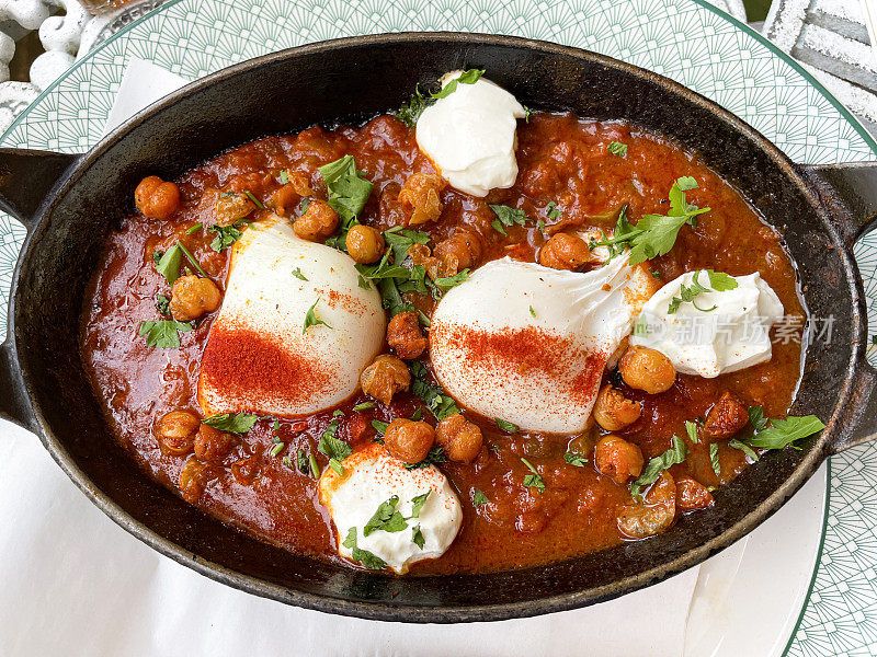 一锅地中海健康早餐的图片，shakshouka / shakshuka / chakchouka放在桌上的盘子上，鹰嘴豆，鸡蛋在番茄，辣椒，洋葱，橄榄油和大蒜的酱汁中水煮，上面放着香菜，俯瞰